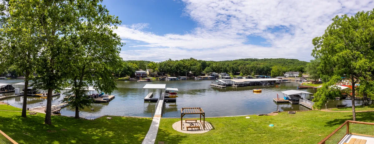 This image shows a peaceful lakeside scene with a view of the lake, docks, lush green trees, and a cozy seating area. The area is clearly set up for activities like boating, fishing, and relaxing by the water, inviting families to interact with the lake just steps from their home.