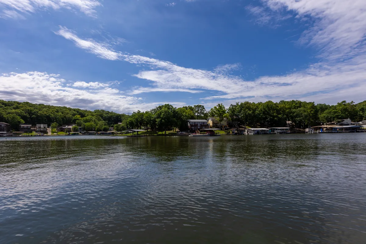 A serene view of the calm lake, framed by lush green trees and houses along the shoreline under a blue sky. The expansive water and greenery offer a visual escape from urban life.