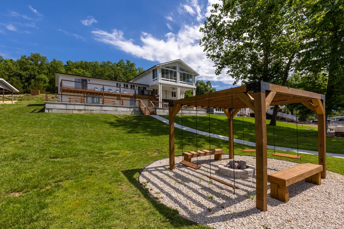 This exterior shot captures the modern house with large windows, a wooden swing set, and a fire pit on a lush green lawn. The space is designed for family gatherings, outdoor play for children, and relaxing evenings by the fire.