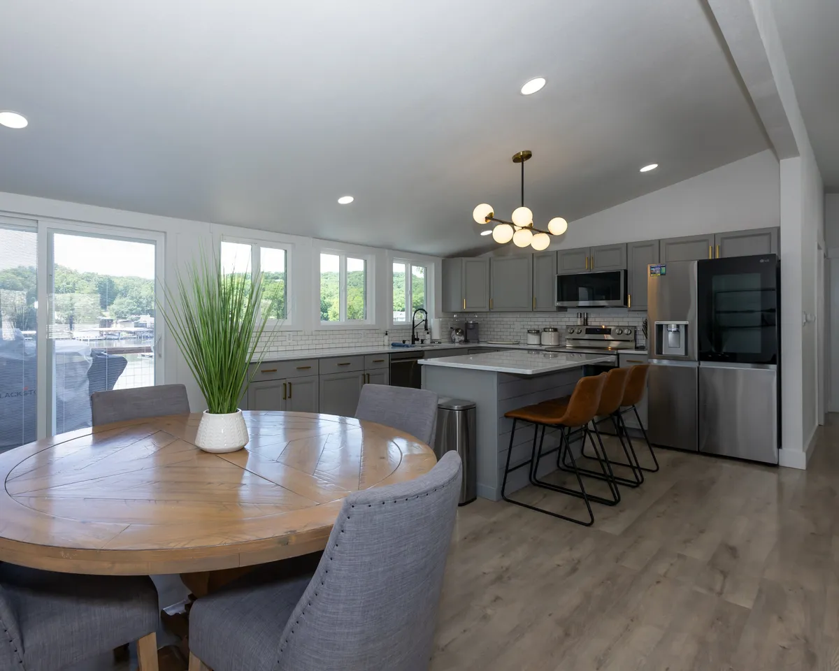 A warm, inviting dining area centered around a round wooden table, illuminated by a modern light fixture. The space includes stainless steel appliances in the background and large windows offering scenic views, reinforcing a communal atmosphere.