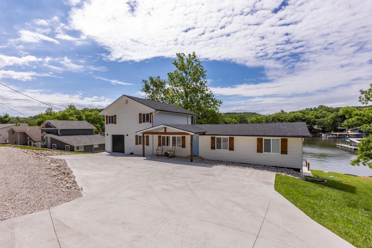 The image features the exterior of the house, including the porch overlooking both the lush greenery and the lake. It highlights the proximity of the home to the water, as well as the inviting outdoor living spaces ideal for gathering and relaxing after a day on the lake.