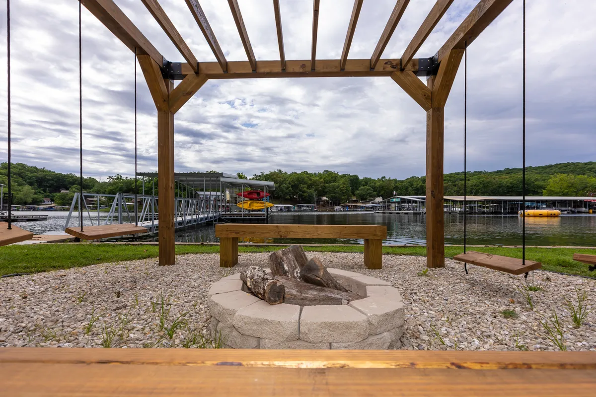 A welcoming exterior scene featuring wooden swings, a fire pit, a pier, and panoramic lake views. The layout encourages socializing, relaxing by the fire, or swinging by the water, inviting guests to enjoy both day and night on the lake. The image captures the playful and communal spirit of the space.