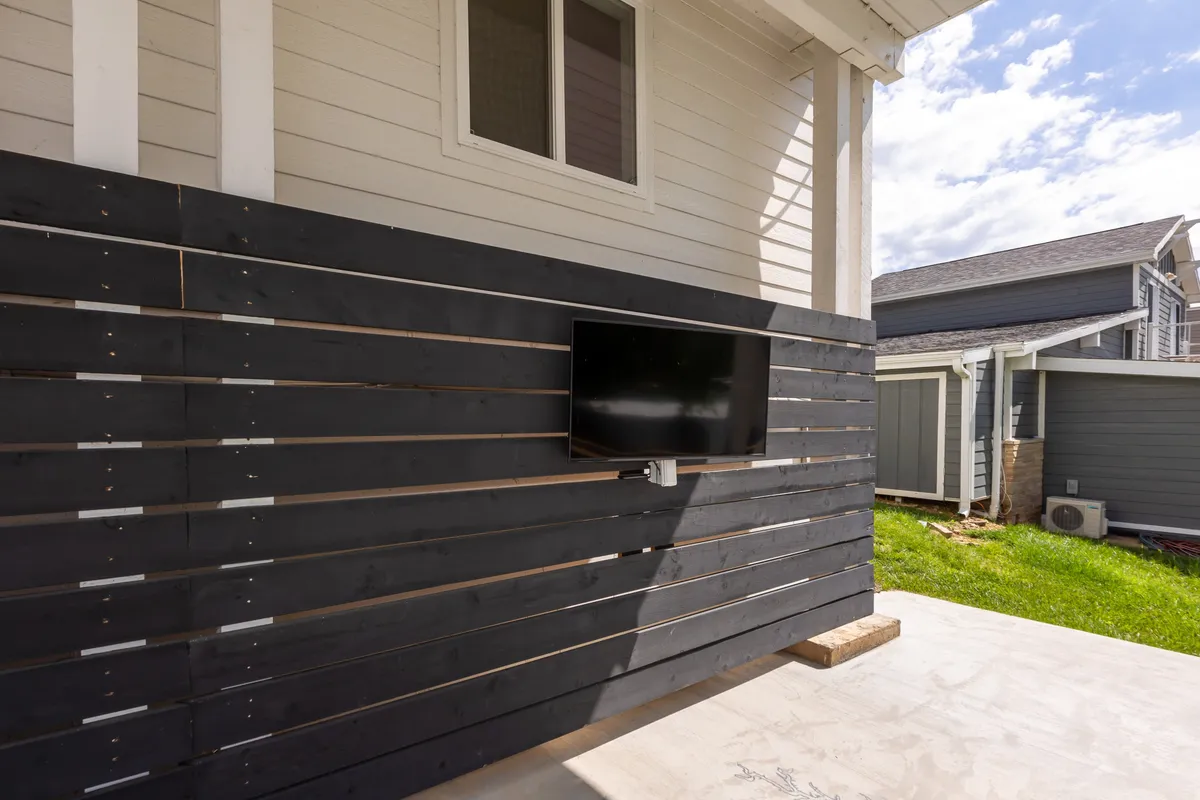 An outdoor space with a modern black wooden wall, a mounted television, and a green lawn. The setting is arranged for relaxing or entertaining, perfect for outdoor movie nights or celebrating special occasions as a group.