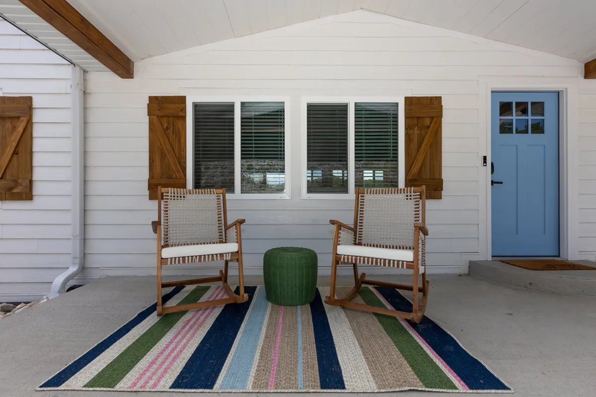 The image shows a welcoming front porch area with rocking chairs, a green stool, and a striped rug. The blue front door adds a bright, cheerful touch, and the setting suggests a space for relaxing with coffee or chatting with friends while enjoying the outdoors.