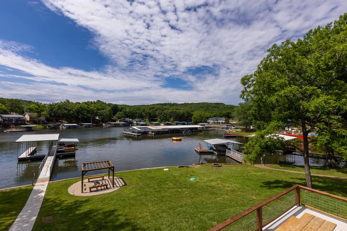 A wide view of the lake with several docks, a green lawn, and a seating area by the water. The image suggests ample space for boating, fishing, and relaxing, with a peaceful vibe enhanced by soft, natural light. The shoreline and water access are both clearly depicted, giving a sense of the property's lakeside amenities.
