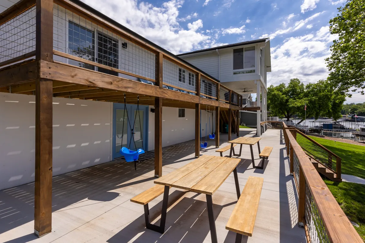A spacious wooden deck overlooking the water, with picnic tables and blue swings set amid lush greenery. The setting provides ample space for outdoor dining, games, and enjoying the lake views and sunsets.