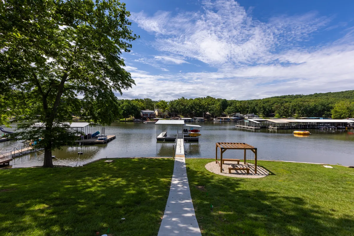 The image captures a dock stretching out into the calm lake, surrounded by lush trees, with a gazebo and boathouses nearby. The dock is clearly visible, inviting activities such as fishing, boating, or simply relaxing by the water. The tranquil setting is enhanced by soft lighting, hinting at either early morning or sunset, and seating is visible for lakeside relaxation.