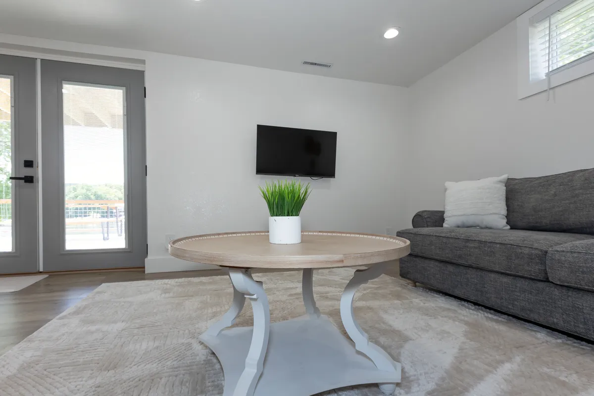 A modern, welcoming living room with a grey sectional sofa, round wooden coffee table, potted plant, wall-mounted TV, and double doors opening to an outdoor deck. The room is filled with natural light, inviting both relaxation and socializing.
