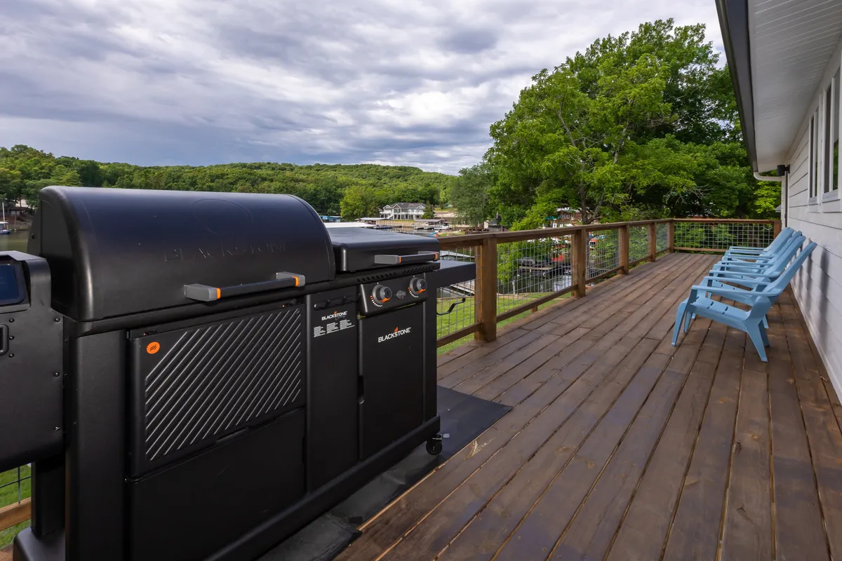 This exterior image shows a well-equipped grill on a deck, blue chairs arranged for socializing, and surrounding green trees. The scene captures an inviting spot for outdoor dining and relaxed gatherings with a scenic backdrop.