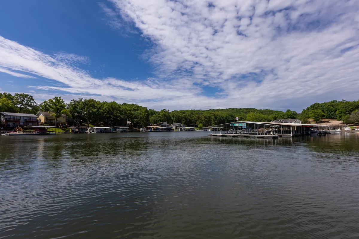 This image displays a tranquil view of calm water with dock structures extending into the lake, surrounded by lush greenery and under a cloudy blue sky. The docks are clearly visible, suggesting direct access for boating and fishing activities, while the peaceful setting hints at relaxation by the water.