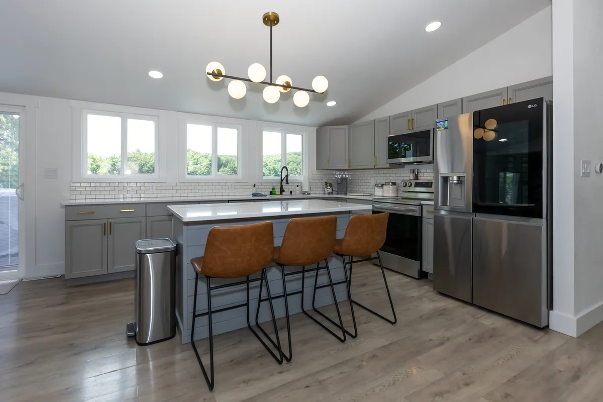 This image features a spacious kitchen with a central island and seating, large windows letting in natural light, a modern light fixture, and stainless steel appliances. The countertops are clear and inviting, perfect for meal prep or casual gatherings.