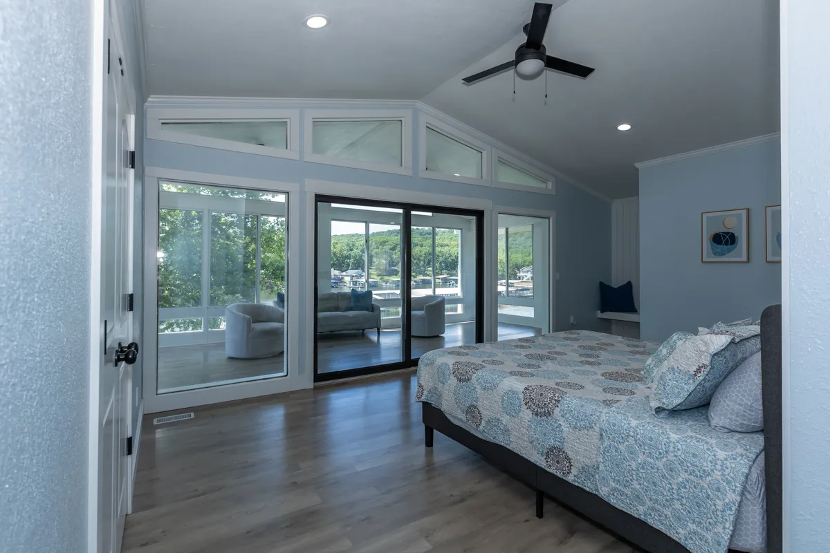 A stylish bedroom featuring a bed with patterned bedding, large windows that offer a beautiful view of the water, and a cozy seating area on the patio just outside. The room is filled with natural light, creating a tranquil and inviting atmosphere.